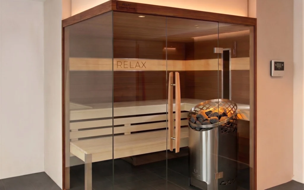 Hochwertige Sauna mit einer Front aus klarem Sicherheitsglas, einer rahmenlosen Glastür und langen Holzgriffen. Im Inneren sind helle Saunabänke vor dunkler Holztäfelung und ein moderner Saunaofen aus Edelstahl sichtbar.