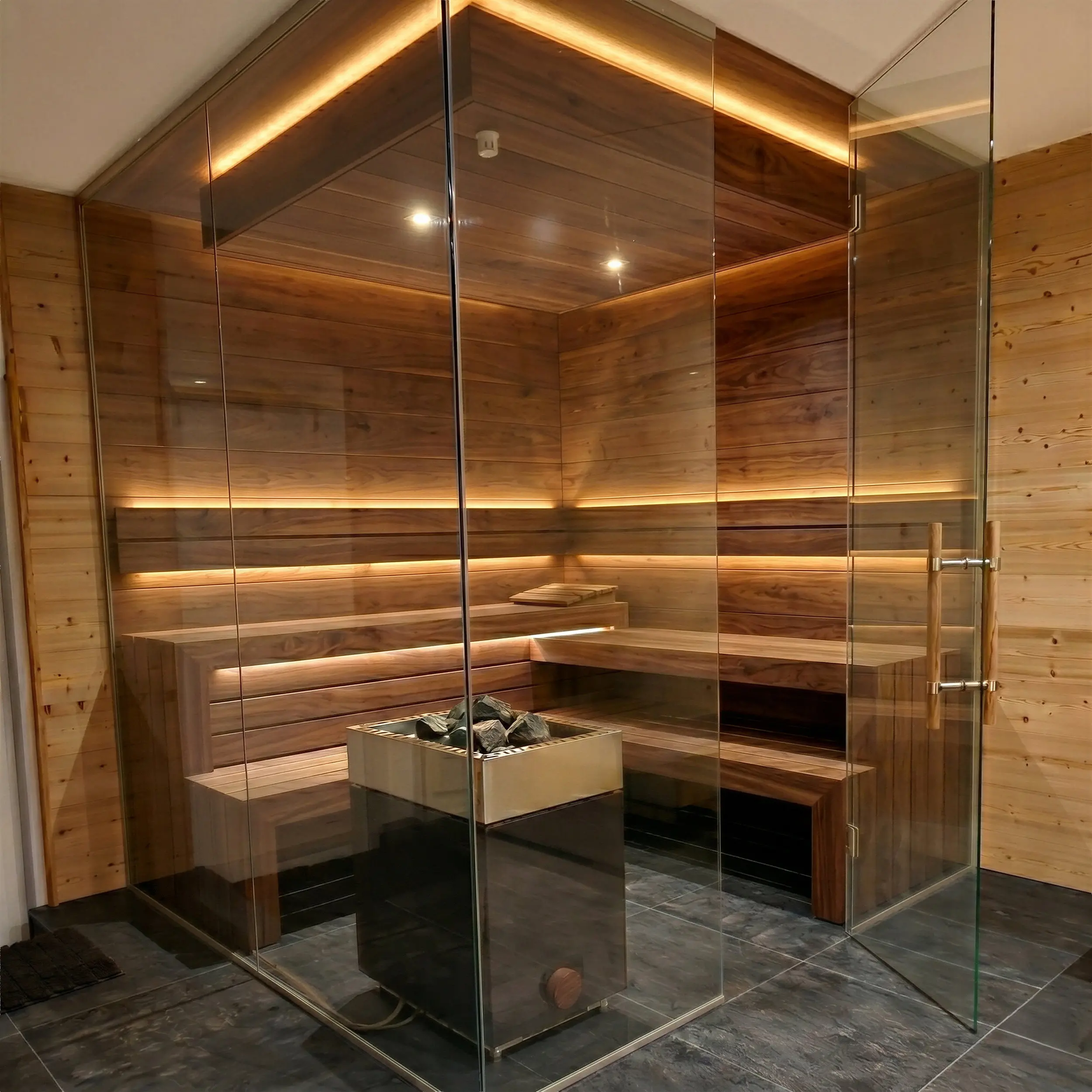 Moderne Design-Sauna mit einer rahmenlosen Eckverglasung aus Glas. Die Ganzglasecke ermöglicht einen transparenten Blick auf die dunkle Holzverkleidung und die beleuchteten Saunabänke. Eine geöffnete Glastür mit Holzgriff führt in den Wellnessbereich.