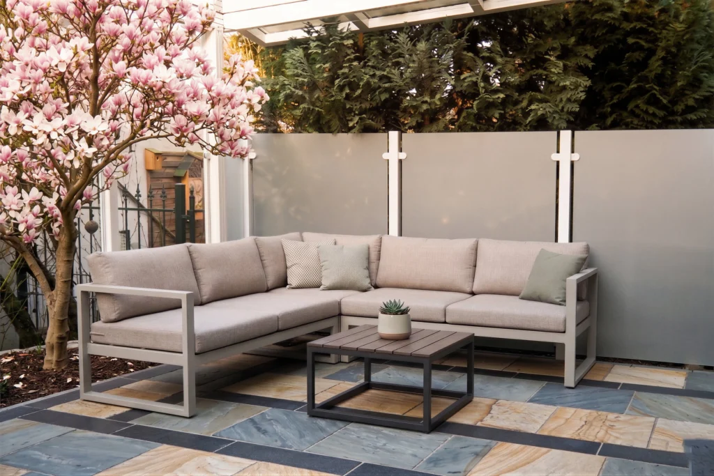 Eine stilvolle Lounge-Ecke auf einer Steinterrasse, abgeschirmt durch einen Sichtschutz aus grau satinierten Glaspaneelen und weißen Pfosten. Neben der Lounge steht eine blühende Magnolie.