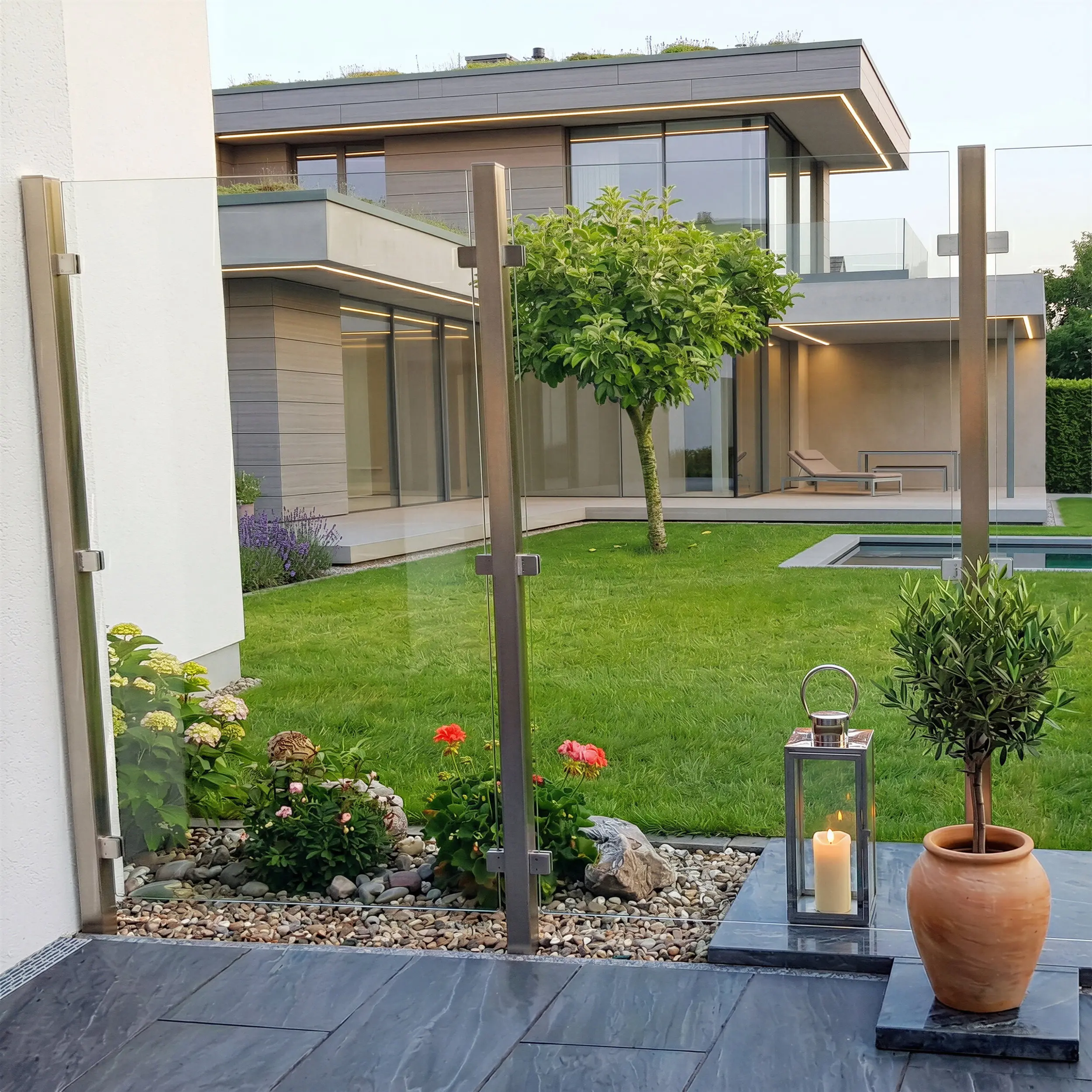 Hochwertiger Glaswindschutz mit stabilen Edelstahlpfosten auf einer dunklen Steinterrasse. Der Blick durch das Klarglas zeigt einen gepflegten Garten und ein luxuriöses, modernes Wohnhaus mit Flachdach und LED-Außenbeleuchtung bei Dämmerung.