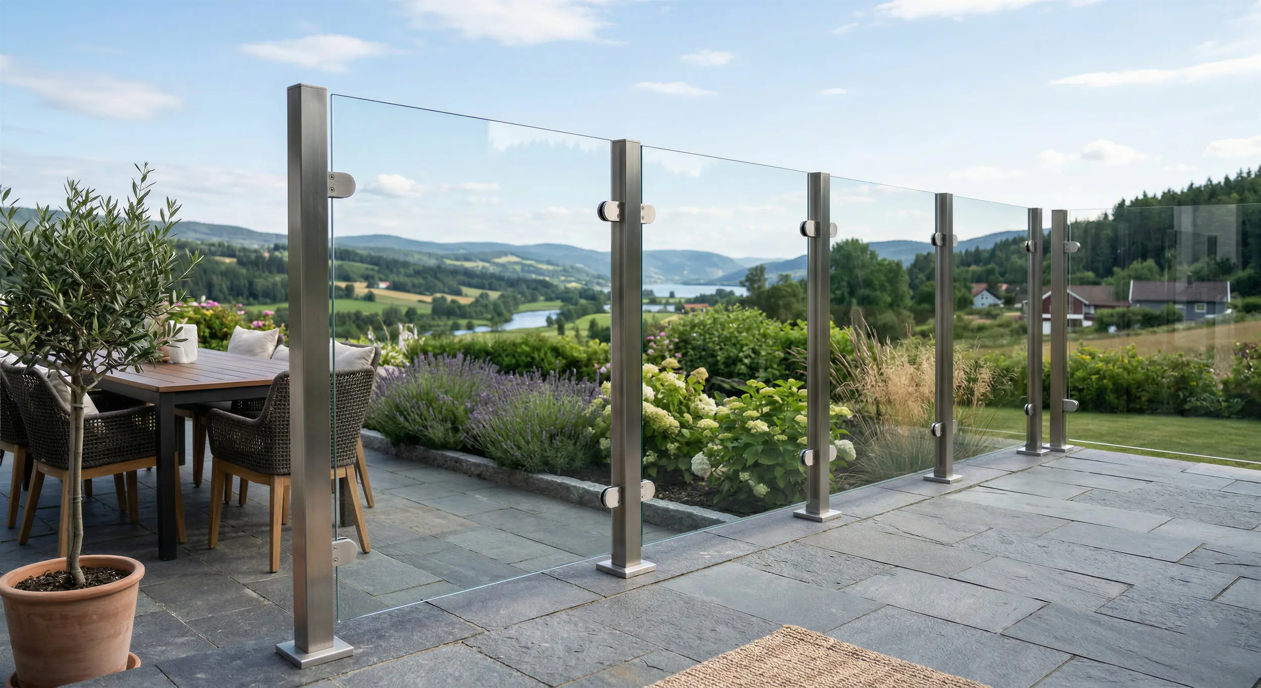 Windschutz-System auf einer Steinterrasse bestehend aus stabilen Edelstahlpfosten, Glasklemmen und klaren Glasscheiben. Im Hintergrund erstreckt sich eine hügelige Landschaft mit einem See und Wäldern.